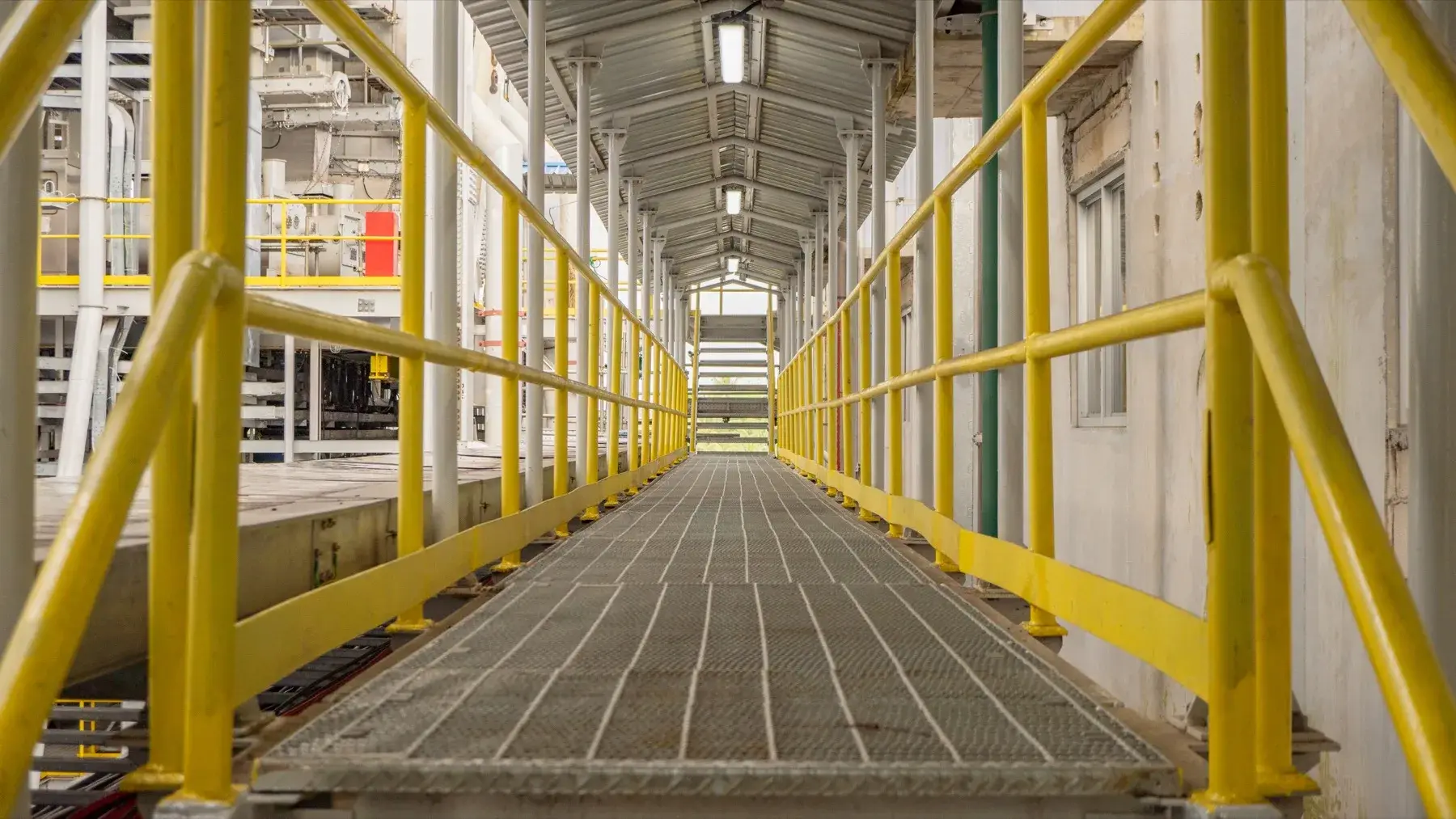 Pasarela elevada de rejilla metálica en una planta industrial, protegida en ambos costados por barandas amarillas de doble travesaño con rodapié
