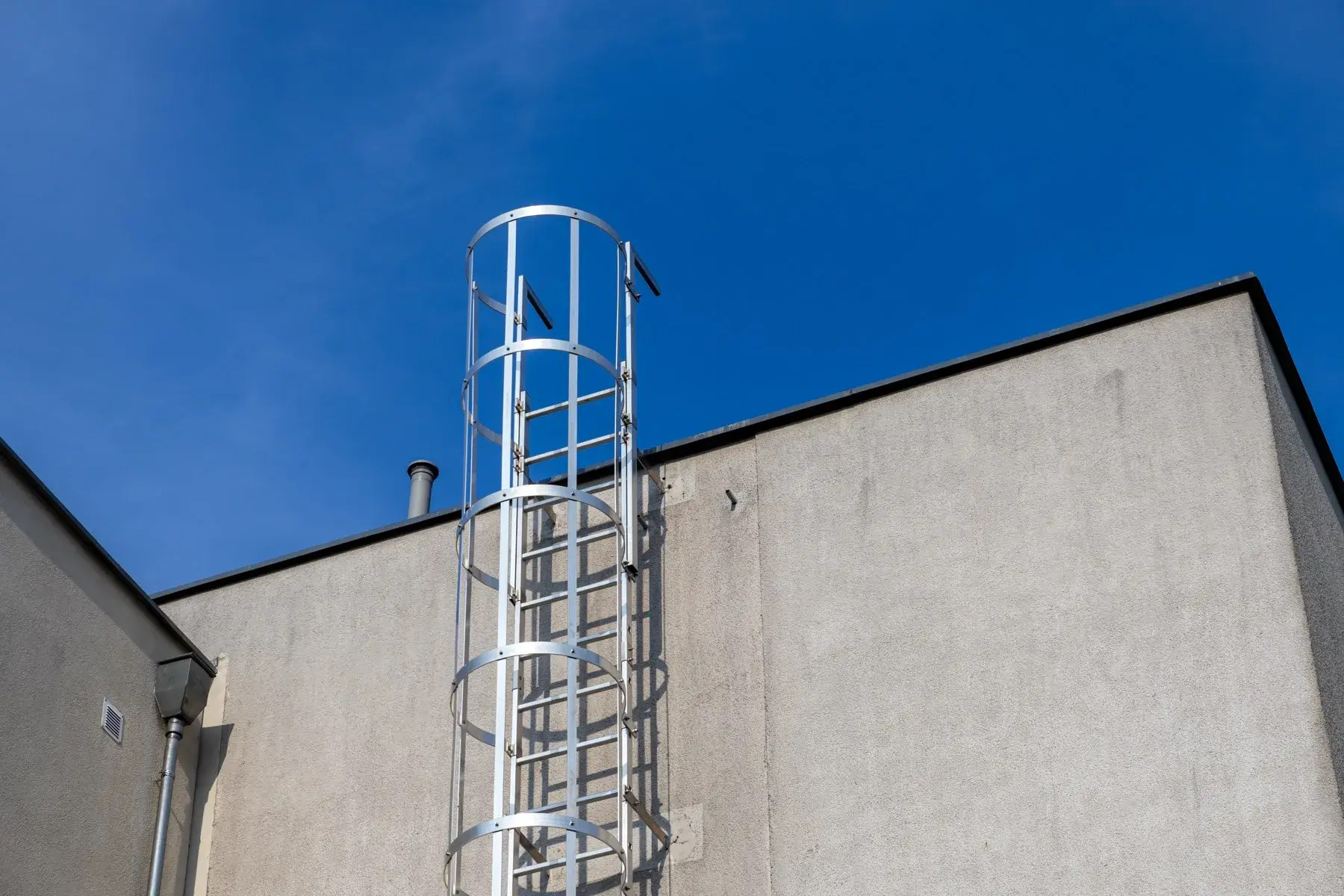 Escala fija vertical de acero galvanizado adosada a la fachada de un edificio, con guarda-cuerpo tipo jaula, plataforma de descanso intermedia y baranda en el nivel de llegada